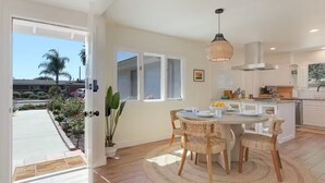A modern kitchen with white cabinets, a dining area with wicker chairs, and a large window overlooking a garden. - Spa, Beaches, Wine Country: Santa Barbara Coastal Escape for Friends and Family (Goleta)