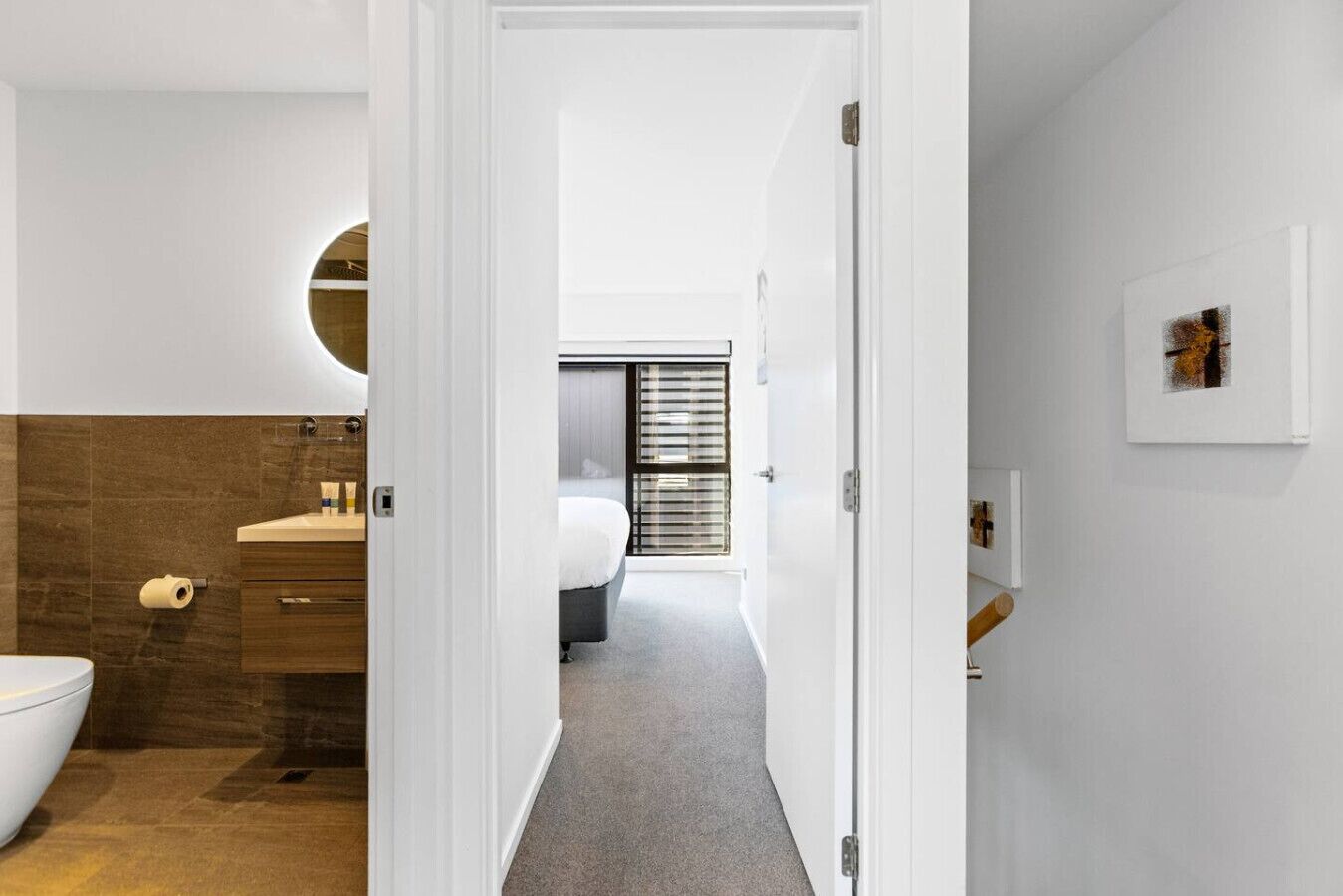 A modern bathroom with a toilet, sink, and shower area. A bedroom with a bed and window blinds is visible through an open door.