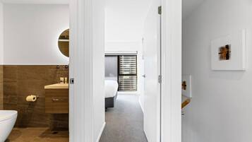 A modern bathroom with a toilet, sink, and shower area. A bedroom with a bed and window blinds is visible through an open door.