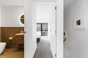 A modern bathroom with a toilet, sink, and shower area. A bedroom with a bed and window blinds is visible through an open door. - Modern Townhouse, Alpine Views, Close To Wanaka (Wānaka)