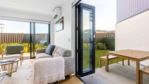 Un salon moderne avec un canapé, une table basse et une table à manger. Une porte-fenêtre coulissante donne accès à un jardin avec une clôture en bois et de la verdure.