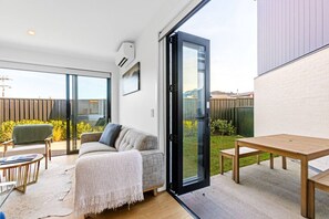 Een moderne woonkamer met een bank, een salontafel en een eettafel. Er is een schuifdeur naar een achtertuin met een houten omheining en begroeiing.