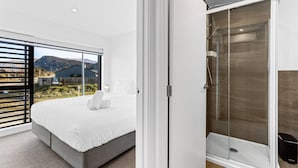 Quarto moderno com cama grande, box de vidro e vista para montanhas e edifícios.