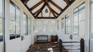 A sunlit room with large windows, wooden beams, and a metal railing.