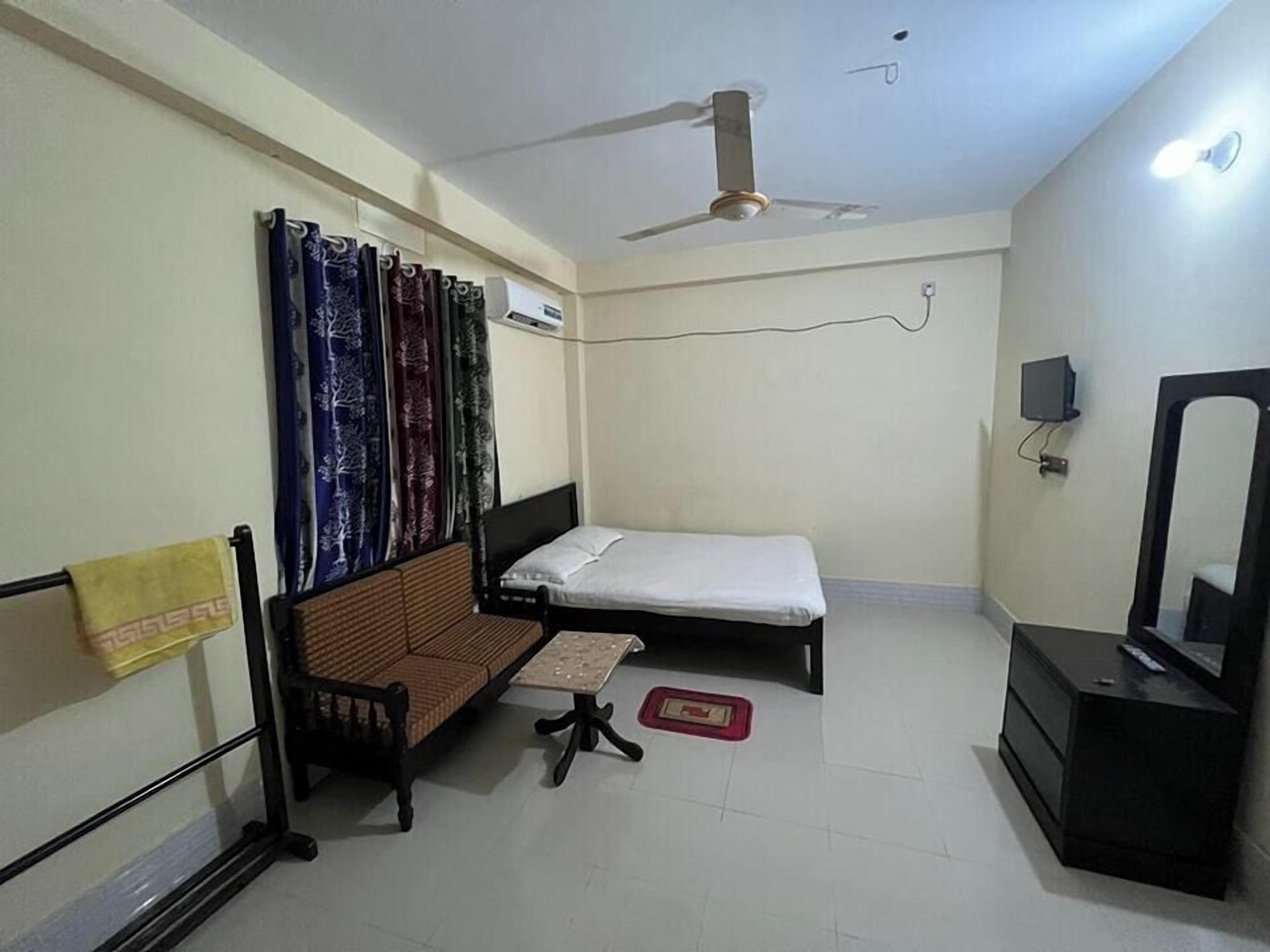 A room with a bed, a sofa, a small table, a mirror, a ceiling fan, and an air conditioning unit.