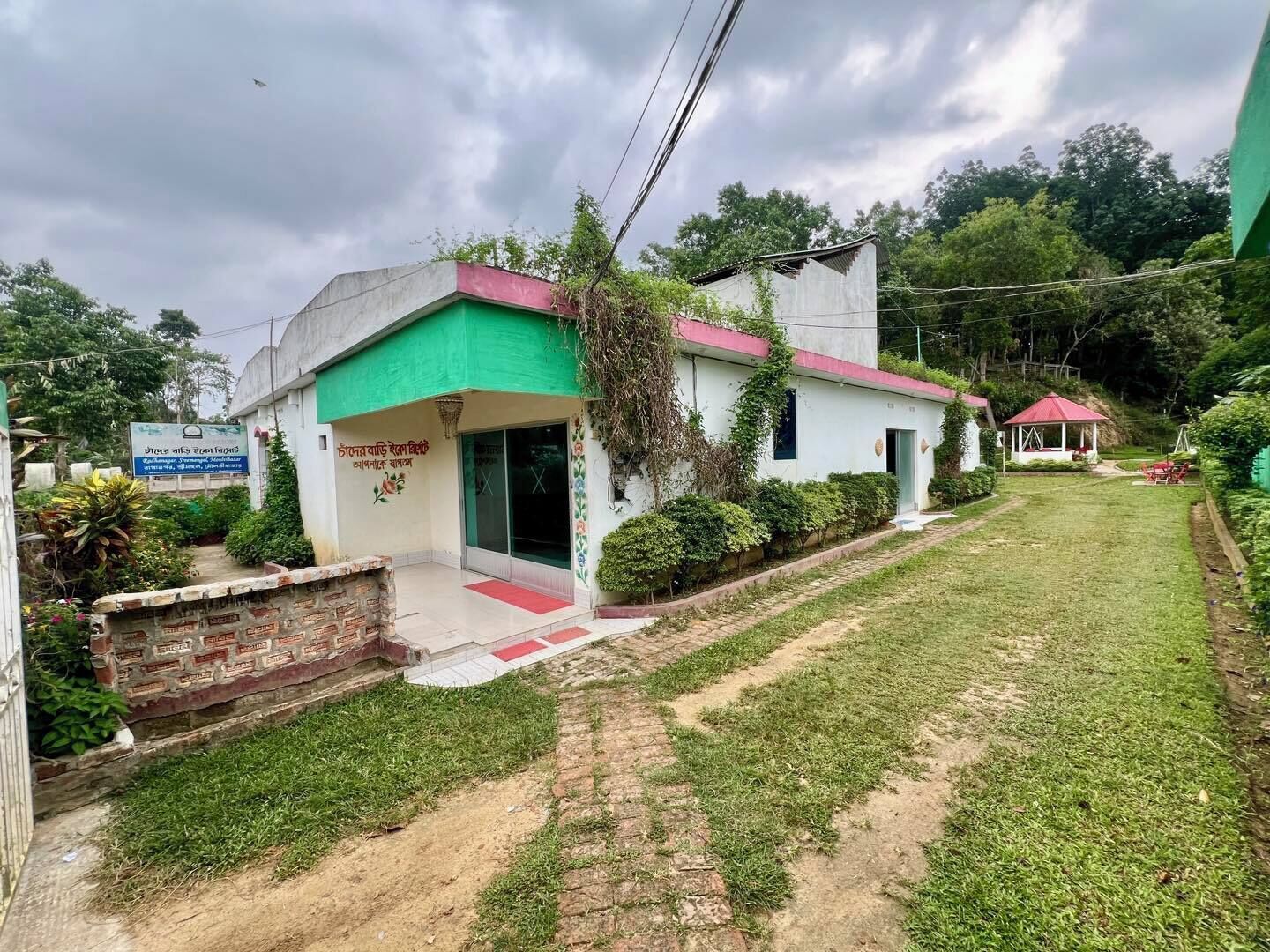 Un edificio de dos pisos con fachada verde y blanca, techo rojo y un pequeño jardín.