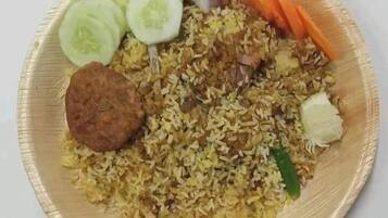 Eine Portion Biryani mit Gurkenscheiben, Karottenstiften und einem Fleischstück.