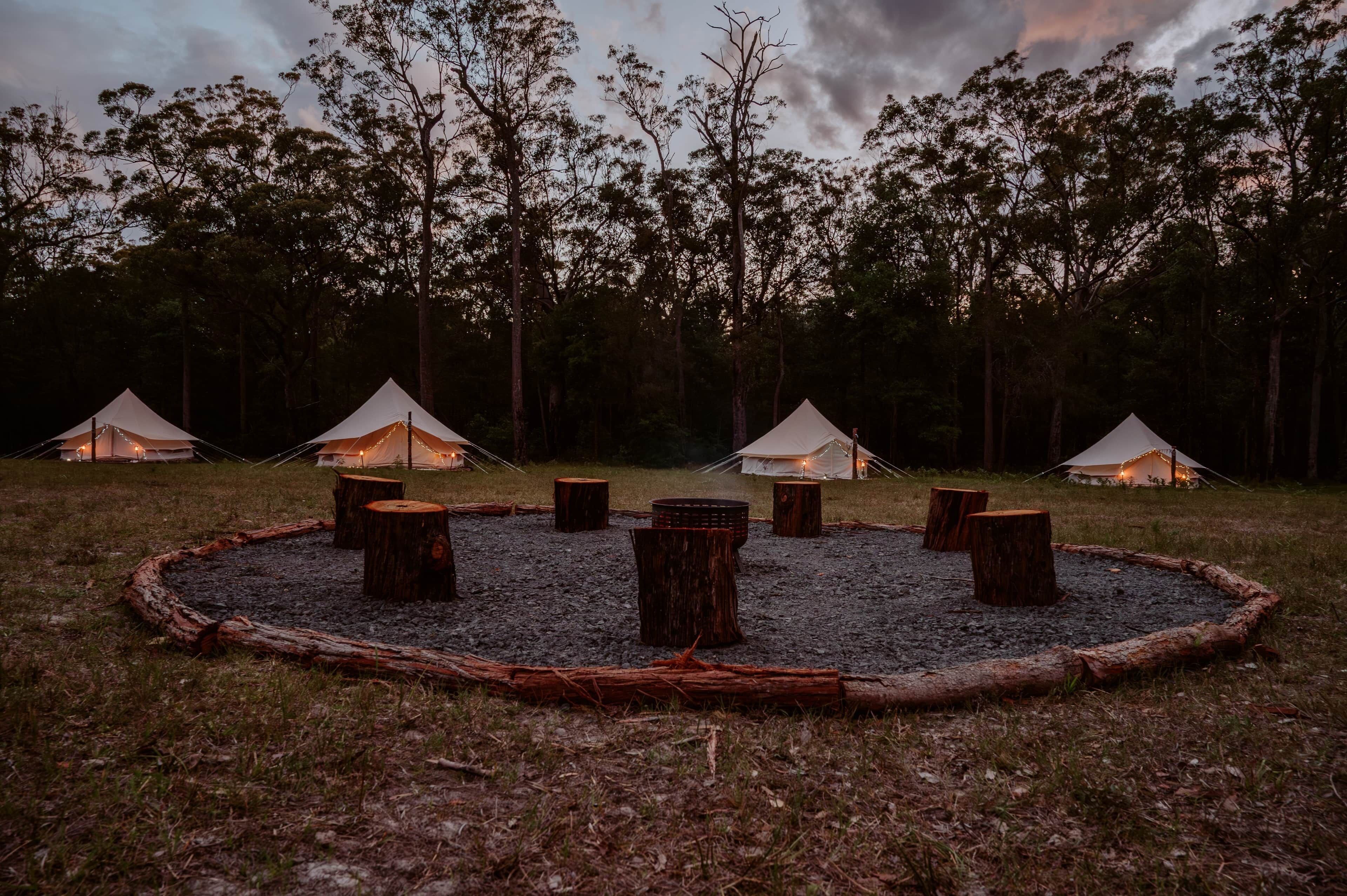 En campingplats med tält som är uppställda i en cirkel runt en central eldstad, omgiven av träd.