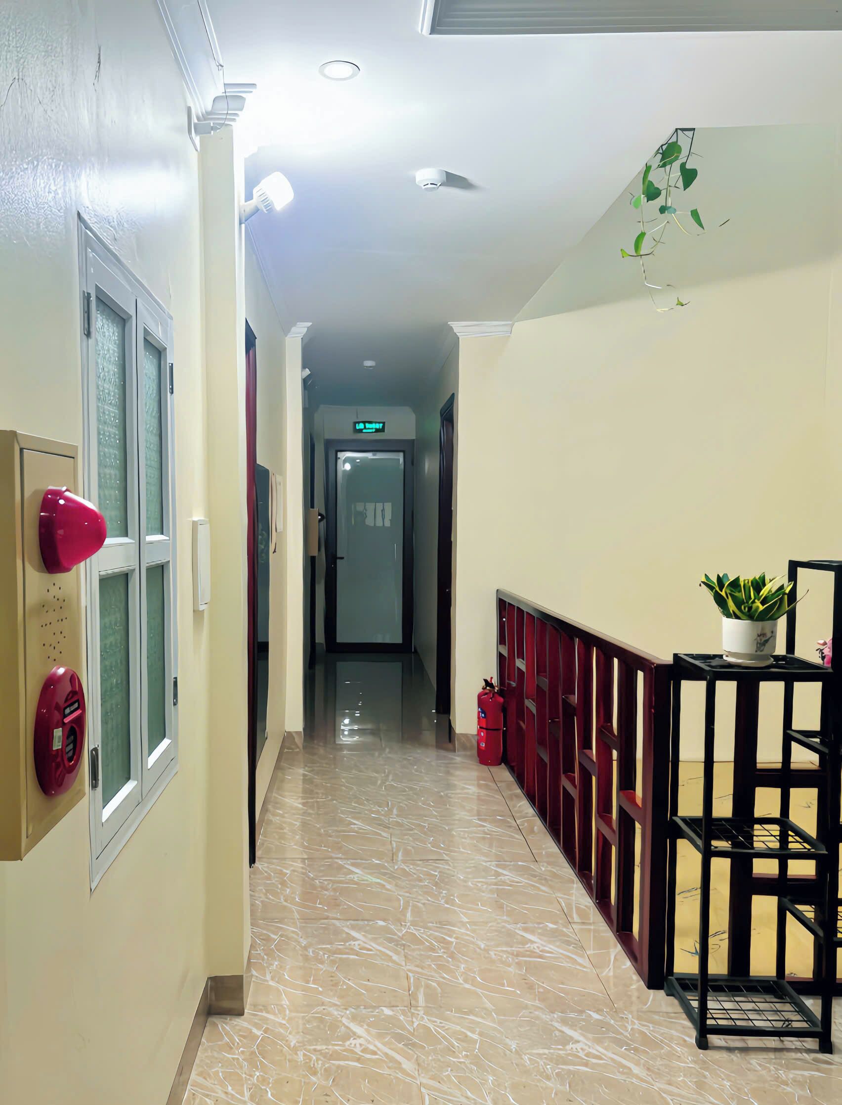 Un couloir d’hôtel avec un sol en marbre, une extincteur rouge, une caméra de surveillance et un pot de fleurs.
