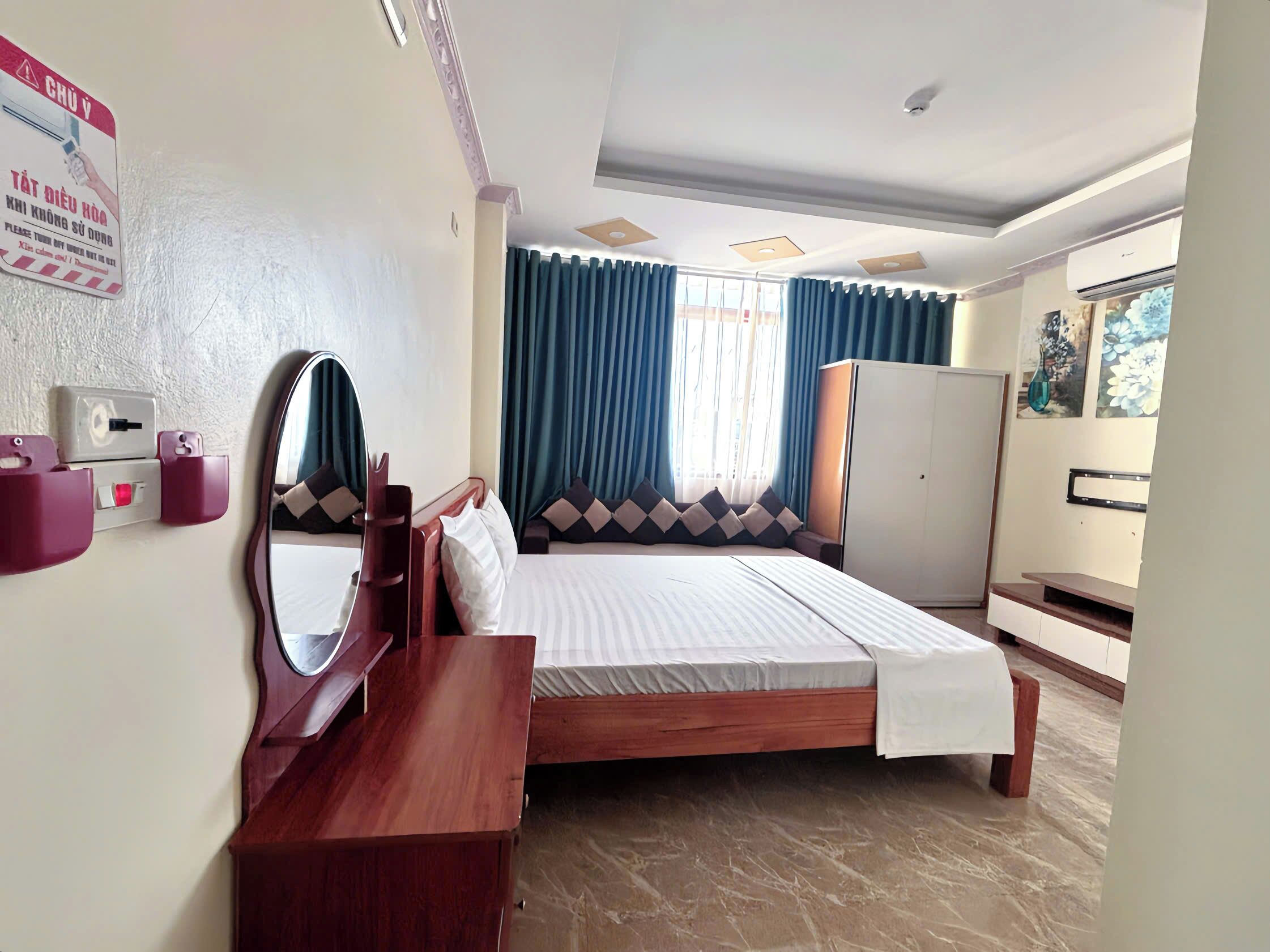 Habitación de hotel con cama, escritorio de madera, armario, espejo y ventana con cortinas.