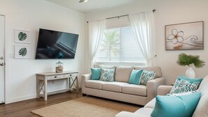 A living room with a beige sofa, a television, a small table, and a window with white curtains.