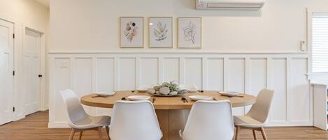A dining area with a round wooden table, white chairs, and three framed botanical prints on the wall.
