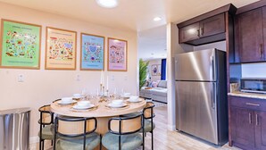Un coin repas avec une table ronde dressée pour six personnes, entourée de chaises en osier. Un réfrigérateur et un four à micro-ondes en acier inoxydable sont visibles dans la cuisine adjacente.