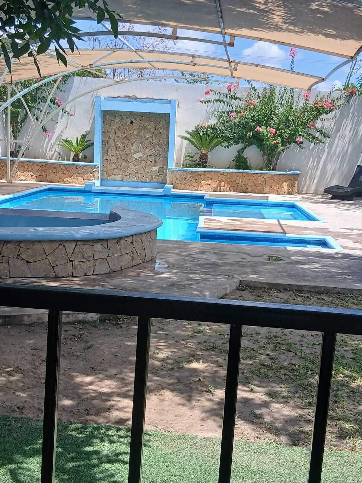 A swimming pool with a stone wall and a small waterfall feature, surrounded by potted plants and a covered seating area.