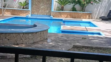A swimming pool with a stone wall and a small waterfall feature, surrounded by potted plants and a covered seating area.