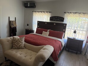 A bedroom with a bed, a chair, a nightstand, a lamp, and a wall-mounted air conditioner.