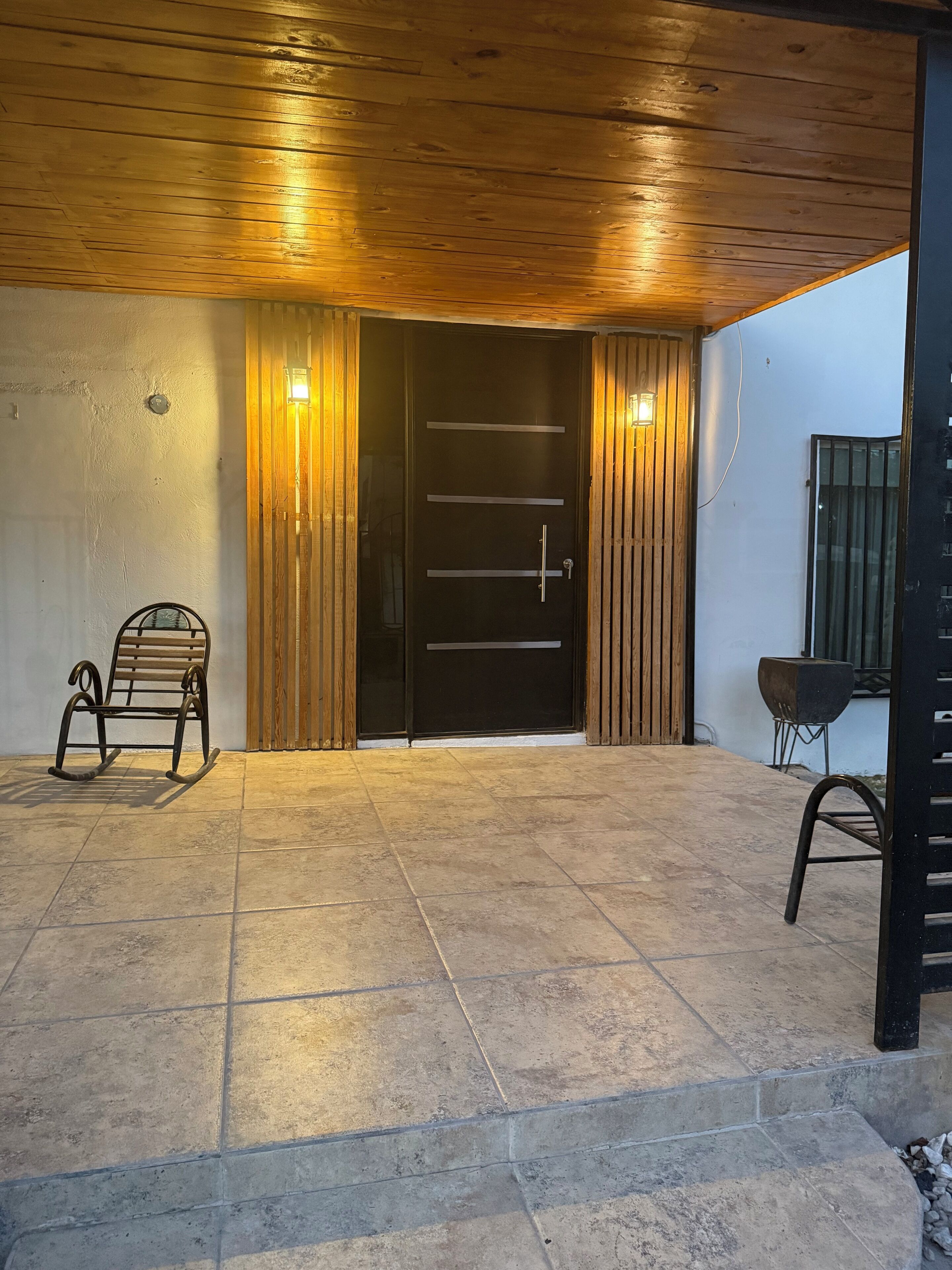 Puerta de madera con un panel negro y una manija, flanqueada por listones de madera verticales y dos luces fijadas a la pared.