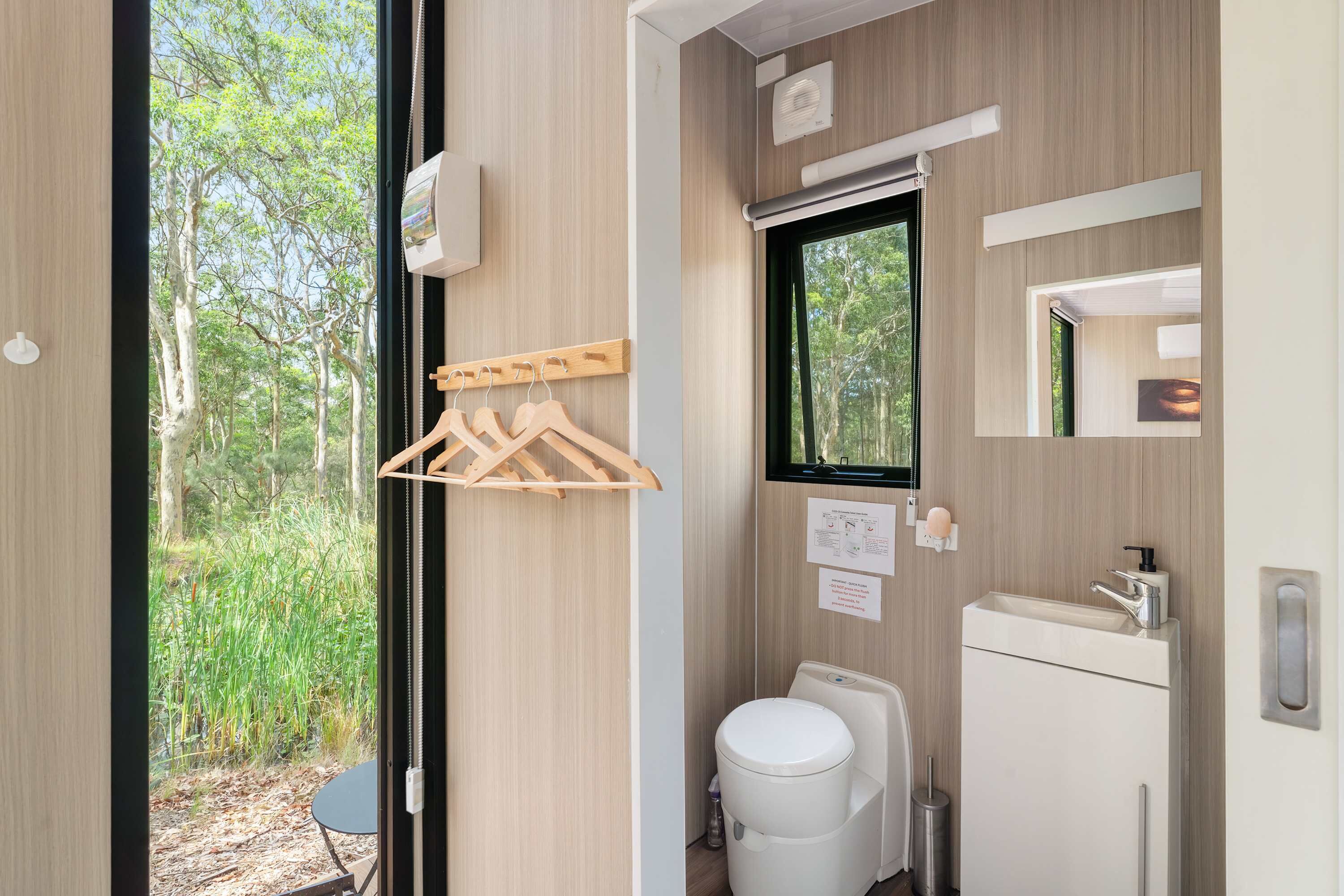 Une salle de bain moderne équipée de toilettes, d’un lavabo et d’un miroir. Une fenêtre laisse entrer la lumière naturelle et offre une vue sur la verdure environnante.