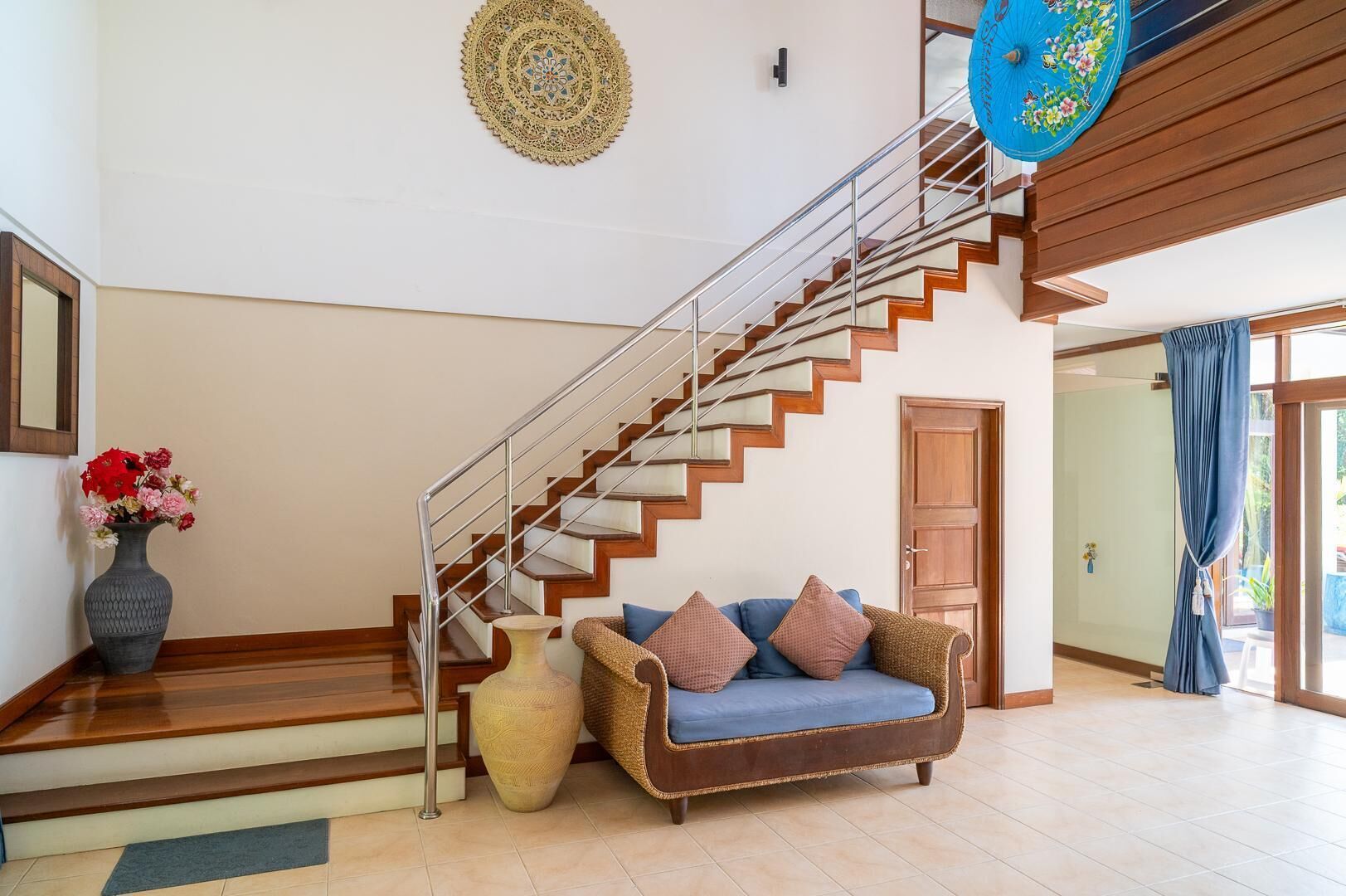 A staircase with wooden steps and metal railings, a woven sofa with cushions, a large vase with flowers, and a decorative ceiling piece.