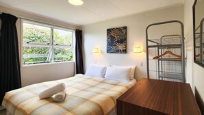 A hotel room with a bed, a wooden desk, a window with curtains, a wall-mounted lamp, and a metal rack with a mirror. - Absolute Beachfront Bach - Classic kiwi bach with absolute beachfront location! (Hahei)