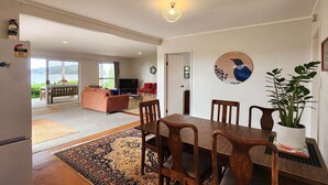 A spacious living area with a dining table, chairs, a sofa, and a television. There is a large window offering a view of a body of water. - Absolute Beachfront Bach - Classic kiwi bach with absolute beachfront location! (Hahei)