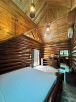 A log cabin interior with a bed, wooden walls, a ceiling light, and a window.