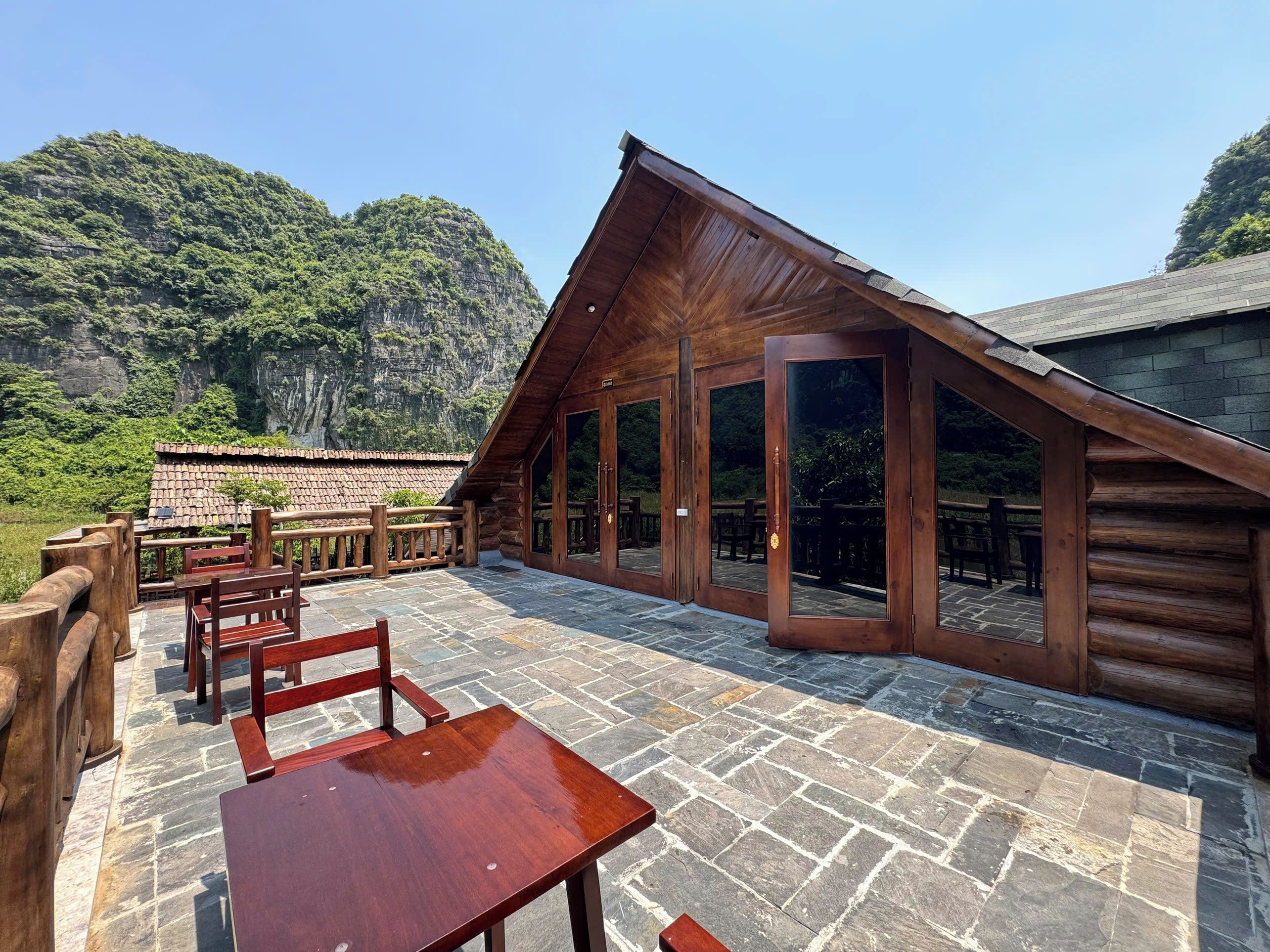 Un edificio de madera con patio de piedra, muebles de madera y vistas a un acantilado boscoso.