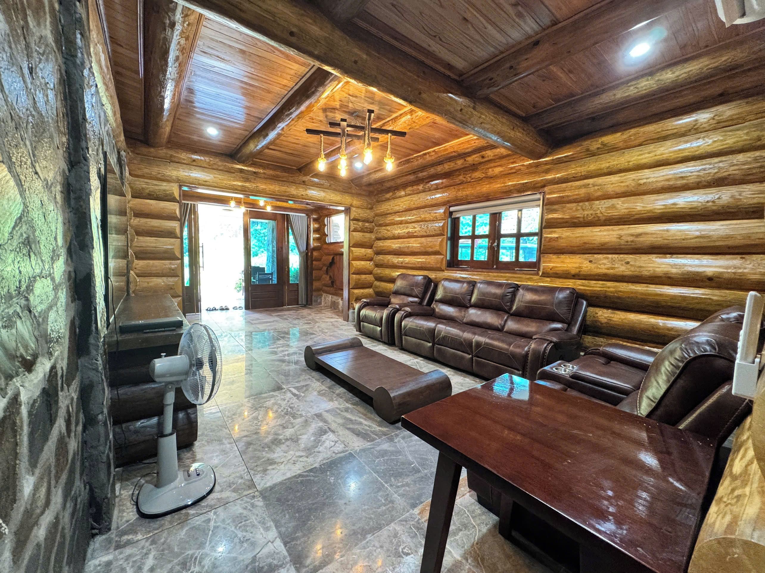 A log cabin interior with a leather sofa, a wooden table, a fan, and a fireplace.