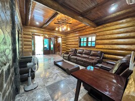 Interior de una cabaña de troncos con un sofá de cuero, una mesa de madera, un ventilador y una chimenea.
