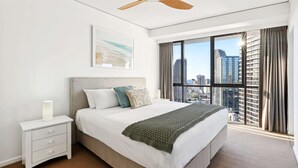 A hotel room with a large bed, a nightstand, a ceiling fan, a window with a city view, and a framed picture on the wall. - Ocean View 3-bed With Gym, Pool (Gold Coast)