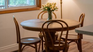 A dining area with a round wooden table, four chairs, a vase with flowers, a floor lamp, and a window with a view of trees. - Charming 2-bedroom cottage in Roscommon (Roscommon)