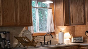 A kitchen with wooden cabinets, a built-in island with bar stools, a coffee maker, and a window with curtains. - Charming 2-bedroom cottage in Roscommon (Roscommon)