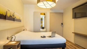 A hotel room with a bed, a desk, a chair, a TV, and a cityscape artwork on the wall. - Ibis Styles Brussels Centre Stephanie (Brussels)