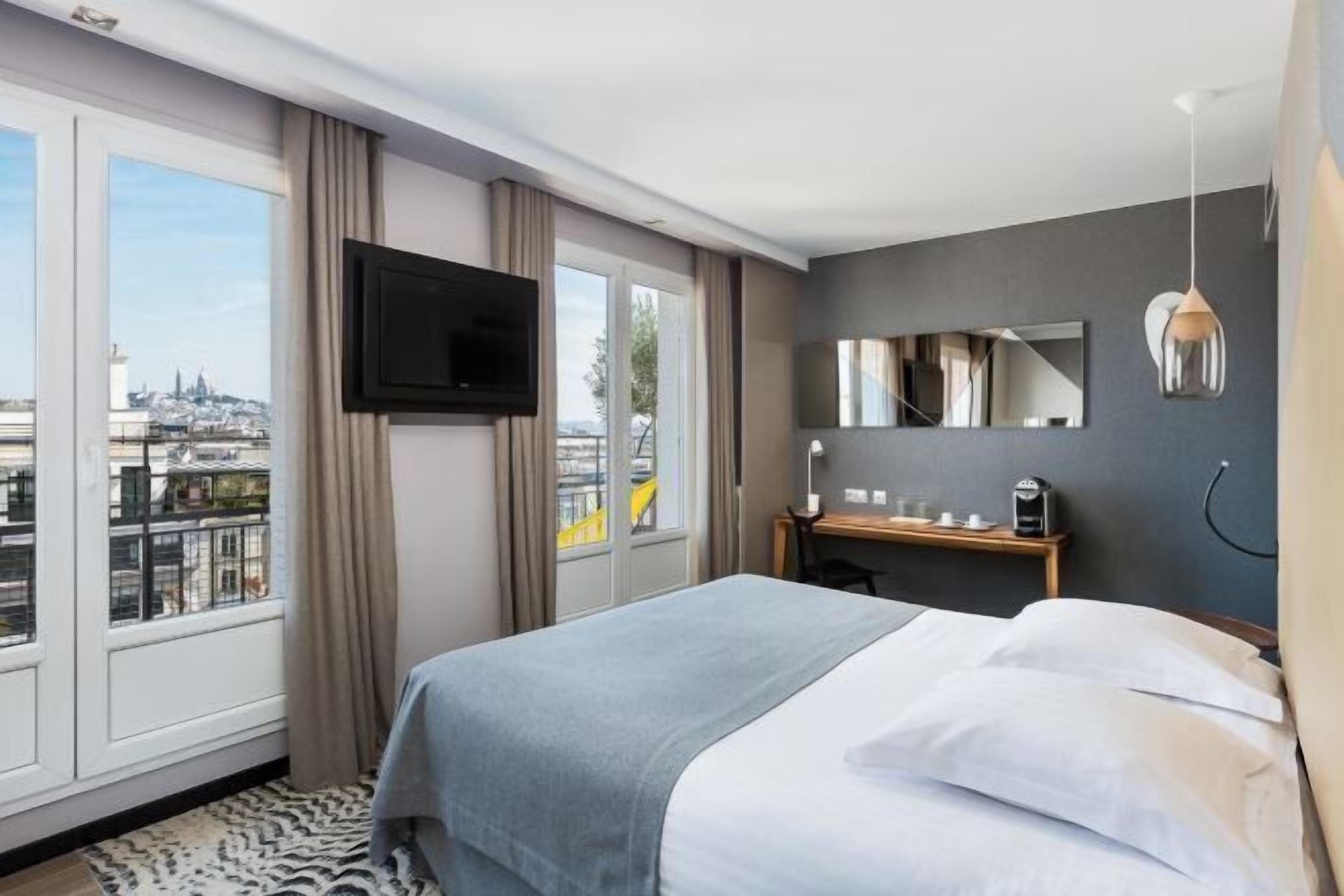 Ein Hotelzimmer mit Bett, Fernseher, Schreibtisch mit Stuhl, Spiegel und Fenster mit Blick auf die Stadt.