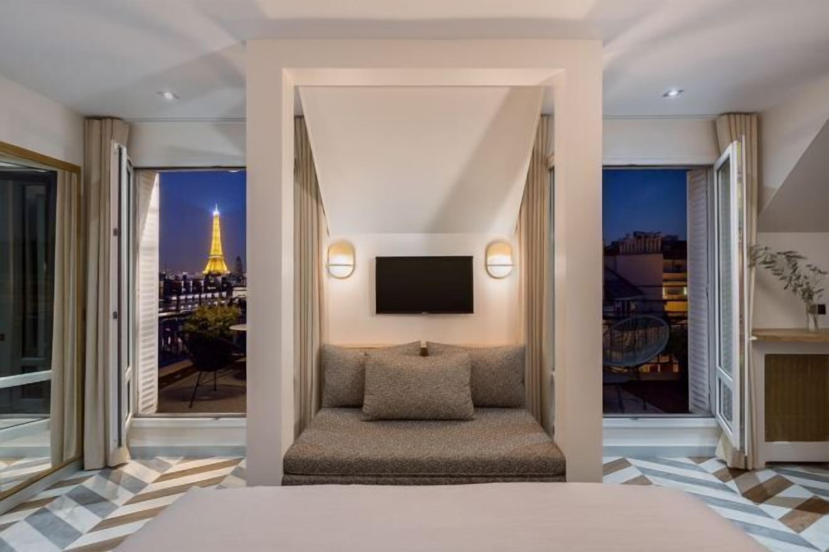 Ein Hotelzimmer mit Bett, Wandfernseher und Blick auf den Eiffelturm bei Nacht.