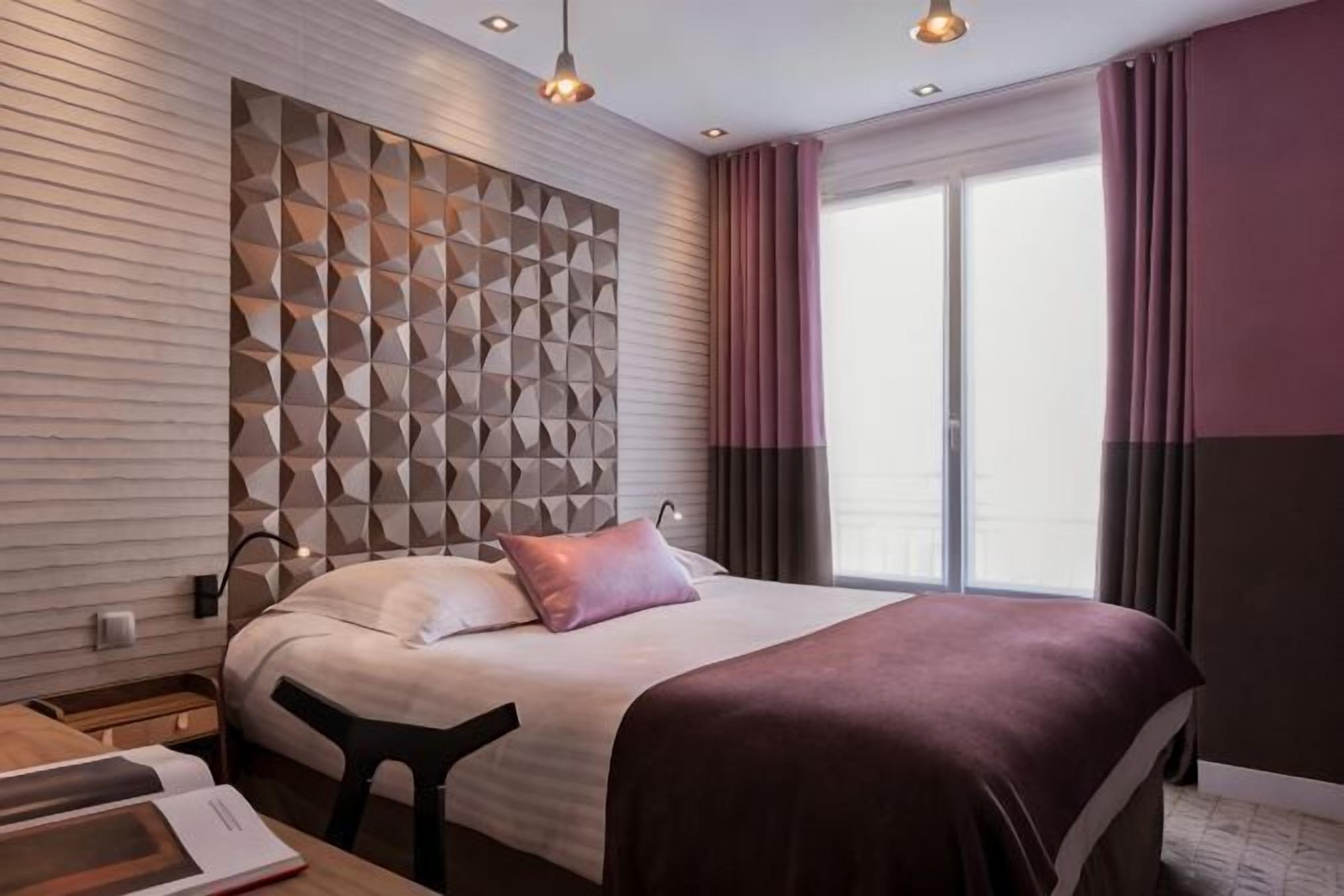 Ein modernes Hotelzimmer mit geometrischem Wanddesign, einem großen Bett mit roter Decke, einem Nachttisch und einem Fenster mit Vorhängen.