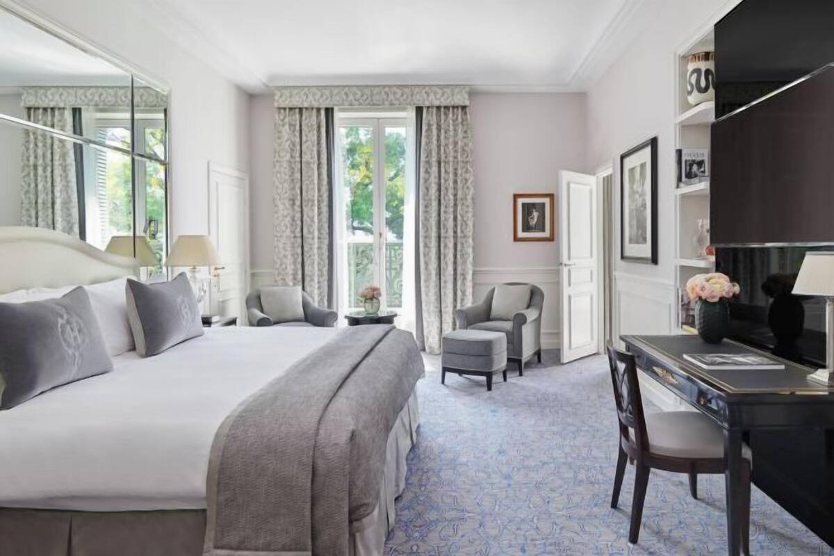 A hotel room with a large bed, a desk, two chairs, a mirror, and a window with curtains.