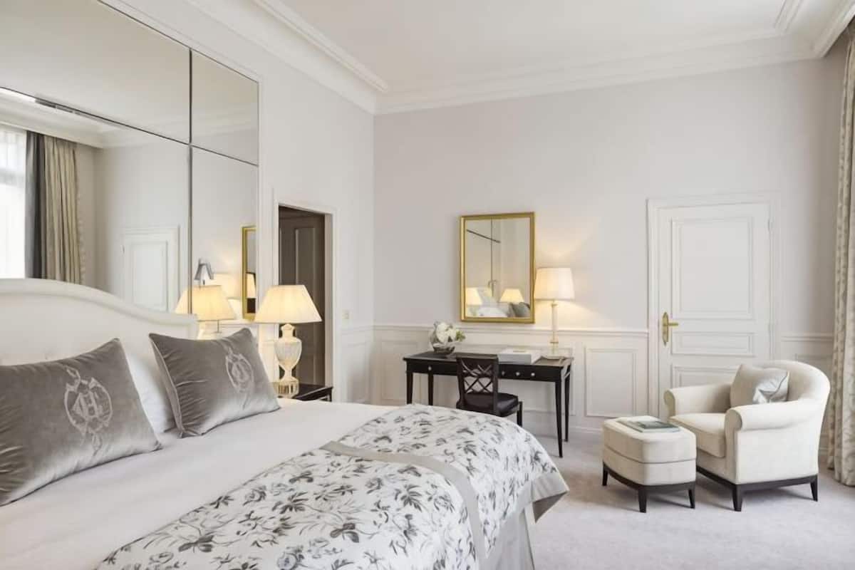 A hotel room with a large bed, a desk, a chair, a small ottoman, and a mirror.