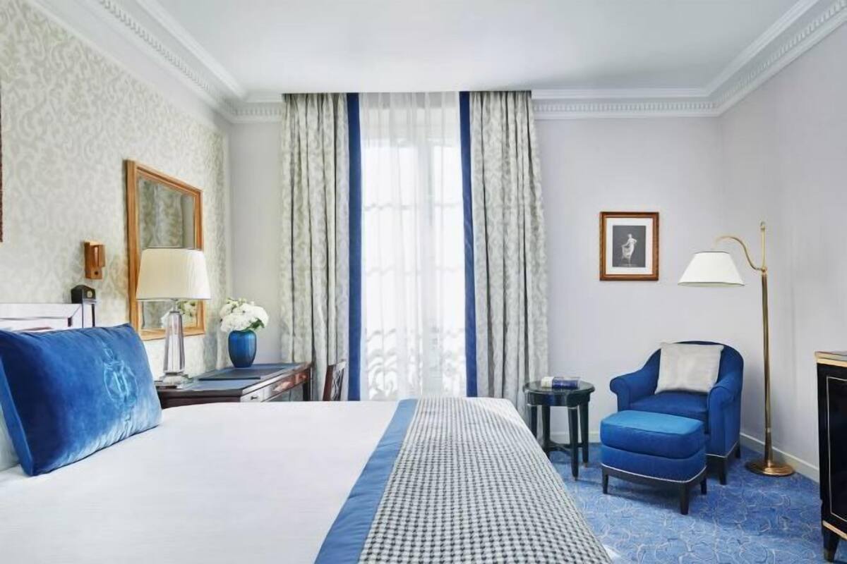 A hotel room with a large bed, a blue armchair, a desk with a lamp, and a picture on the wall.