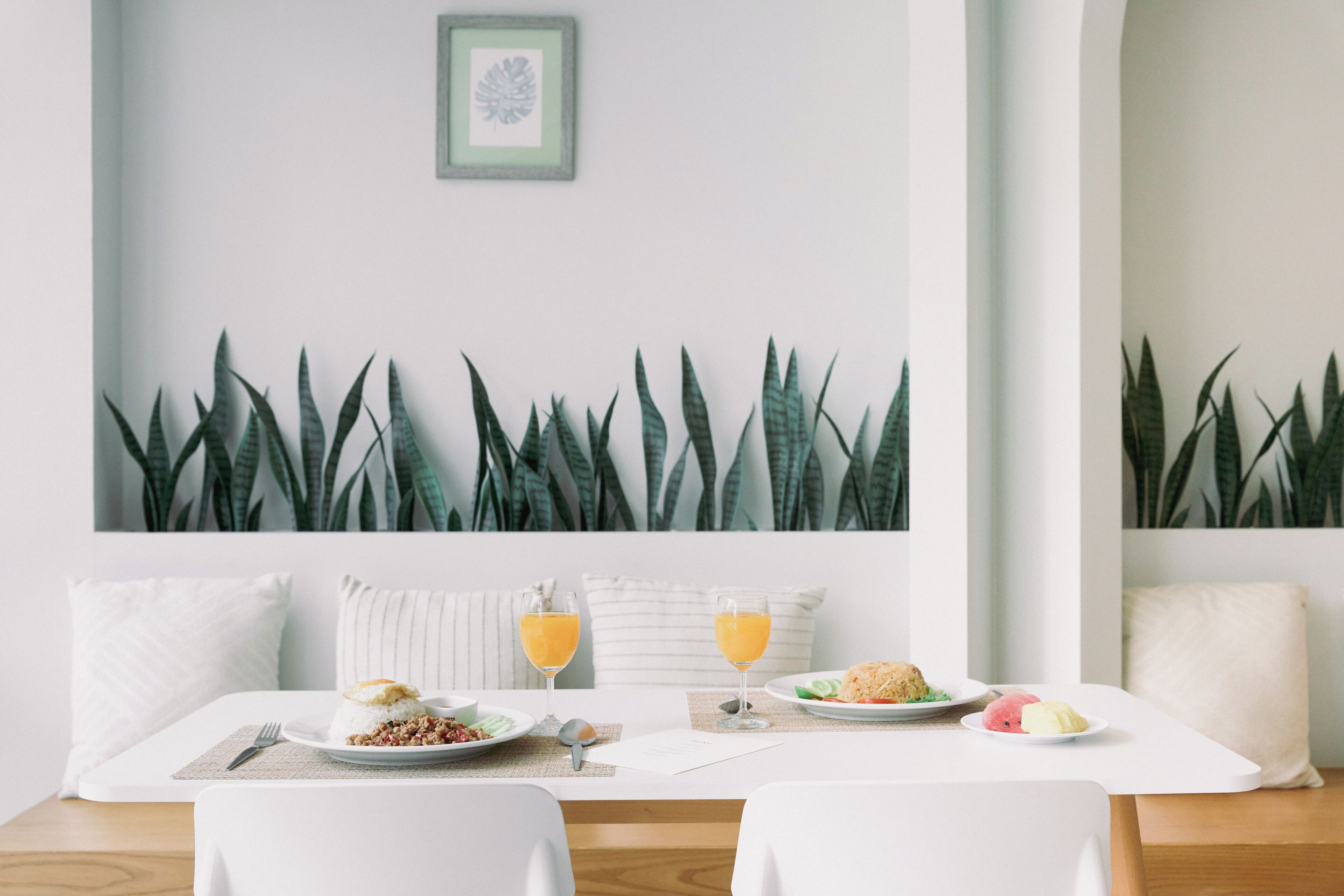 A dining area with a white table, two chairs, a framed picture on the wall, and a row of green plants.