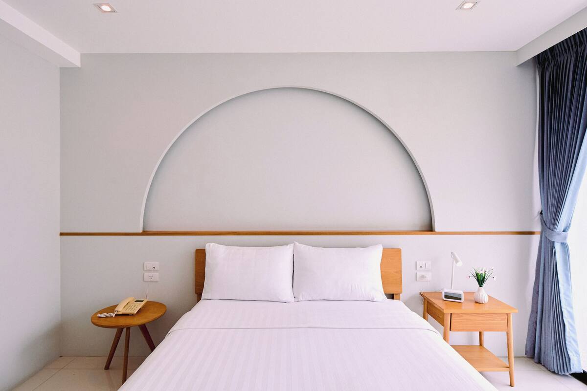 Quarto de hotel moderno com uma cama grande, uma mesa de cabeceira com abajur, uma pequena mesa redonda com telefone e uma janela grande em arco.