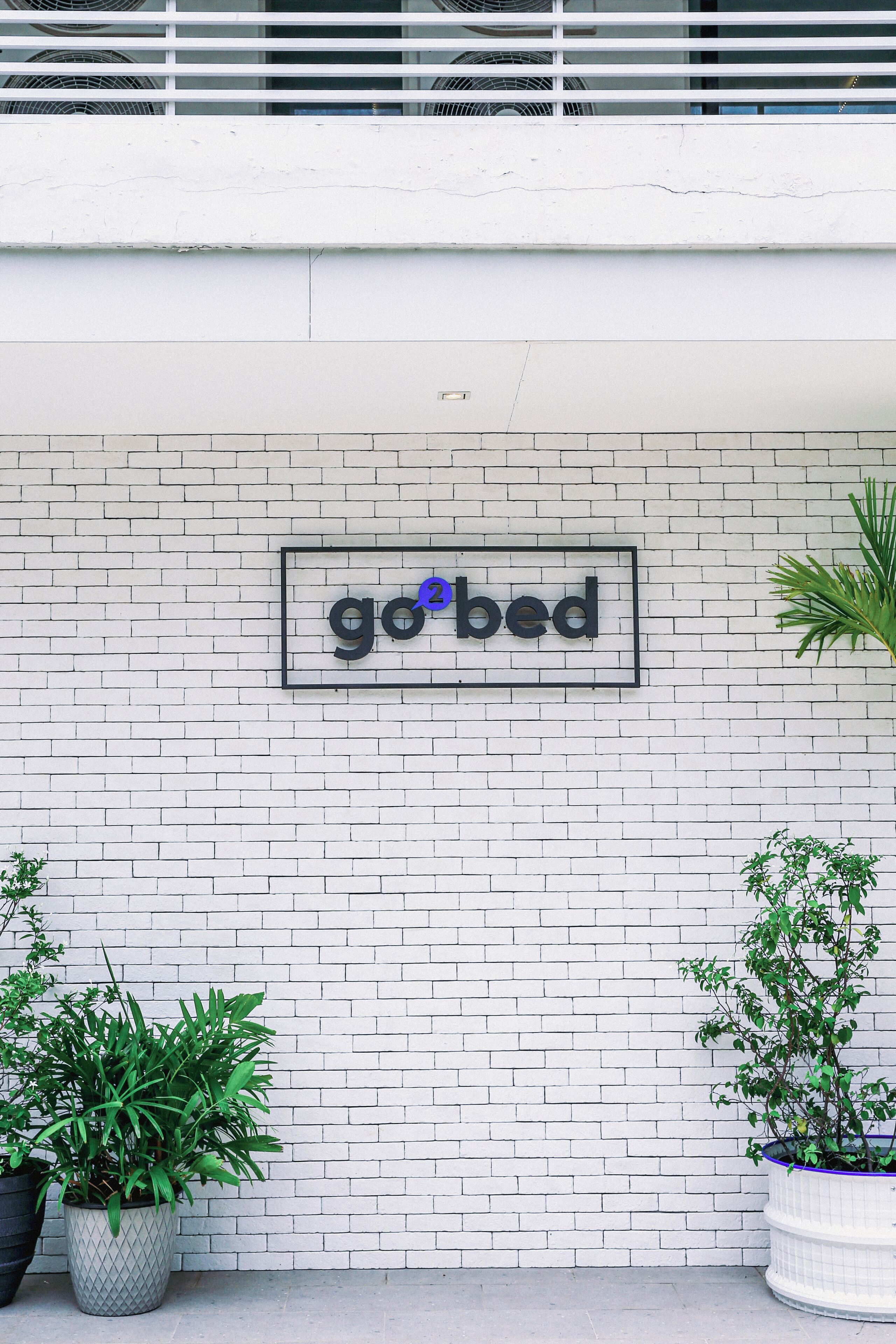A white brick wall with a framed sign that reads "go bed". There are two potted plants on either side of the sign.