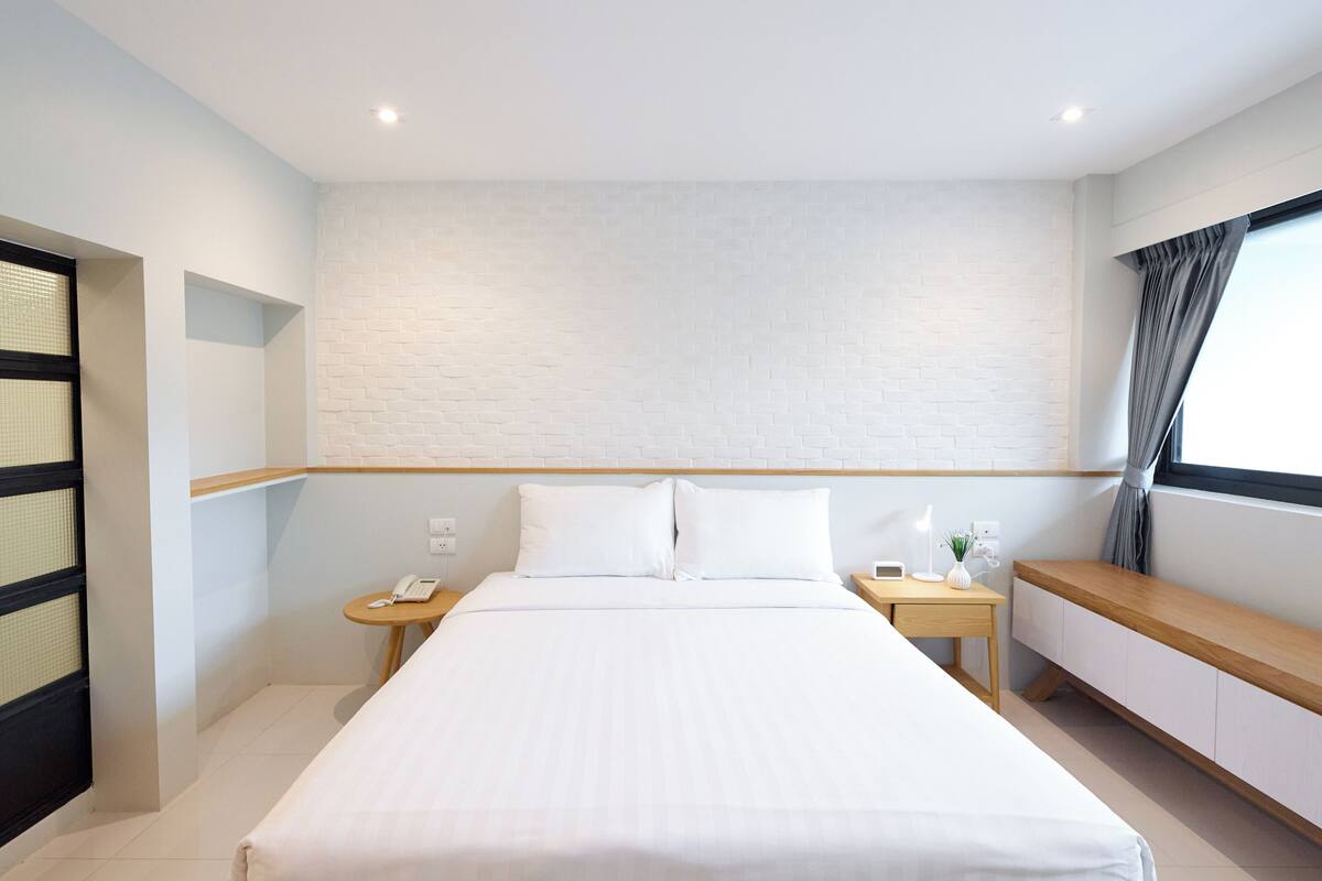 Quarto de hotel com uma cama grande, uma mesa de cabeceira com abajur, uma mesinha com telefone, uma janela com cortinas e uma parede de tijolos brancos.