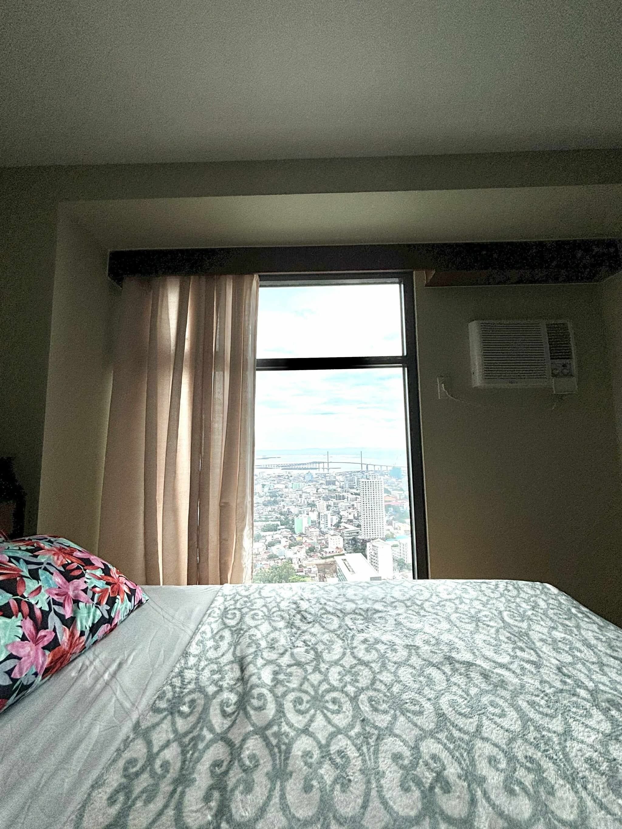 A hotel room with a large bed, a window with curtains, and a view of a cityscape.