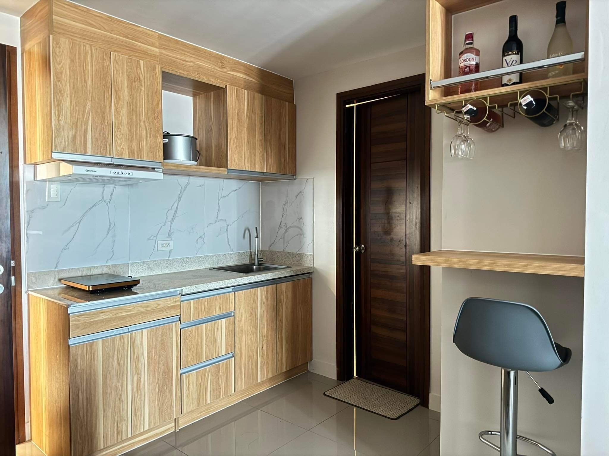 Een moderne keuken met houten kastjes, een roestvrijstalen spoelbak en een inbouwoven. Er is een bar met een stoel en planken vol flessen en glazen.