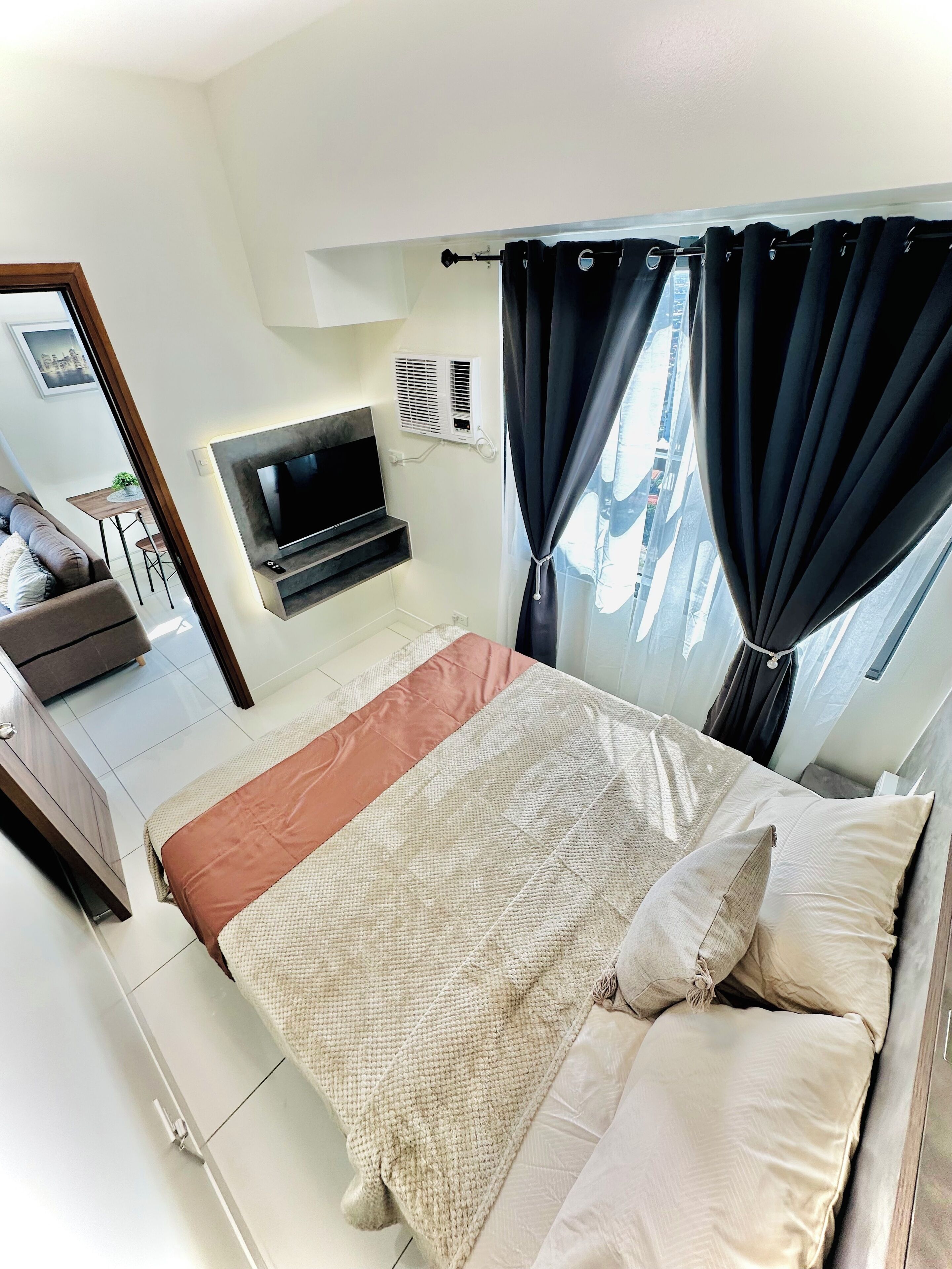 A bedroom with a bed, a TV, a window with curtains, and a small table with a plant.