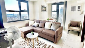 Een moderne woonkamer met een hoekbank, een staande ventilator en uitzicht op de stad door grote ramen.