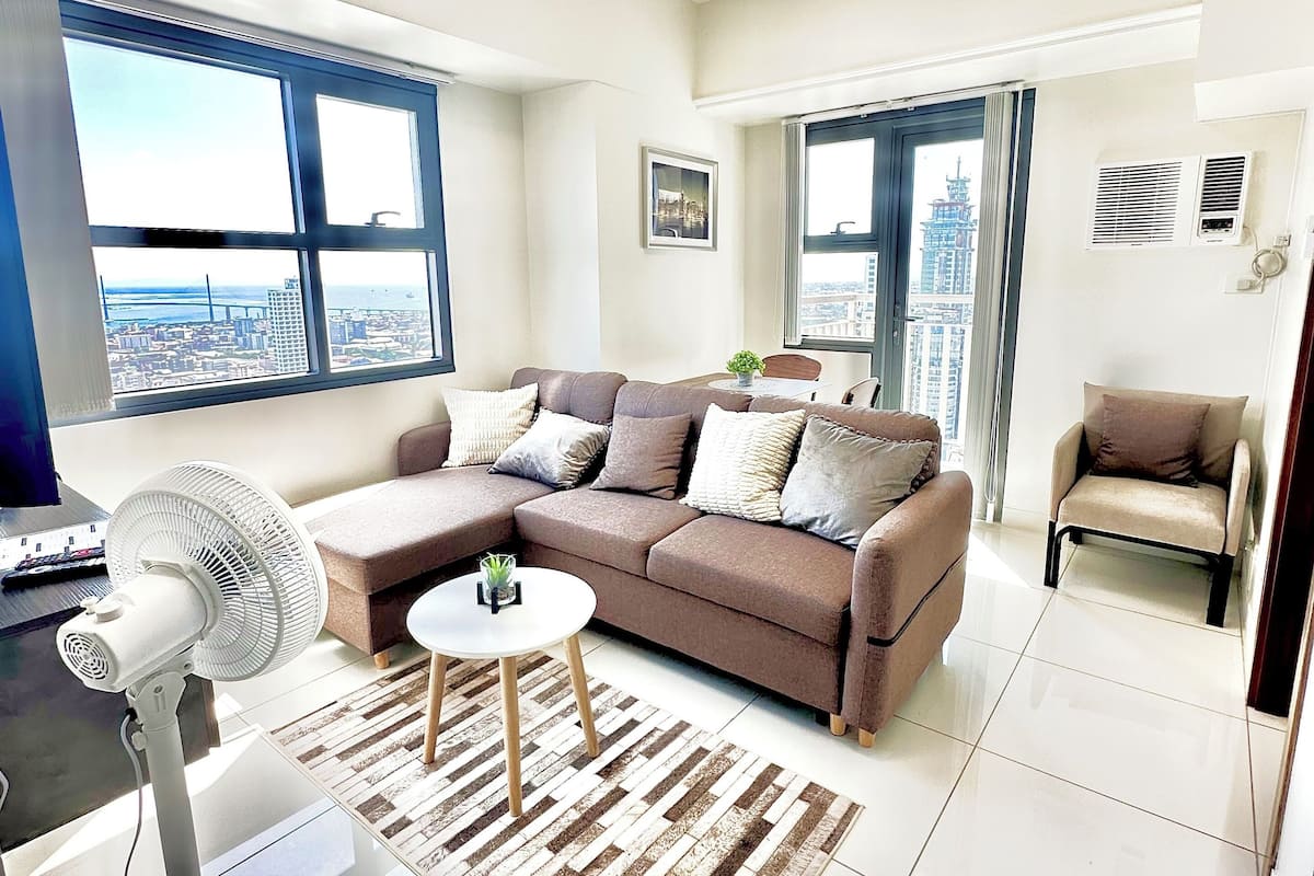 Una sala de estar moderna con un sofá seccional, un ventilador de pie y vistas de la ciudad a través de ventanas amplias.