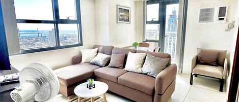 A modern living room with a sectional sofa, a standing fan, and a view of the cityscape through large windows.