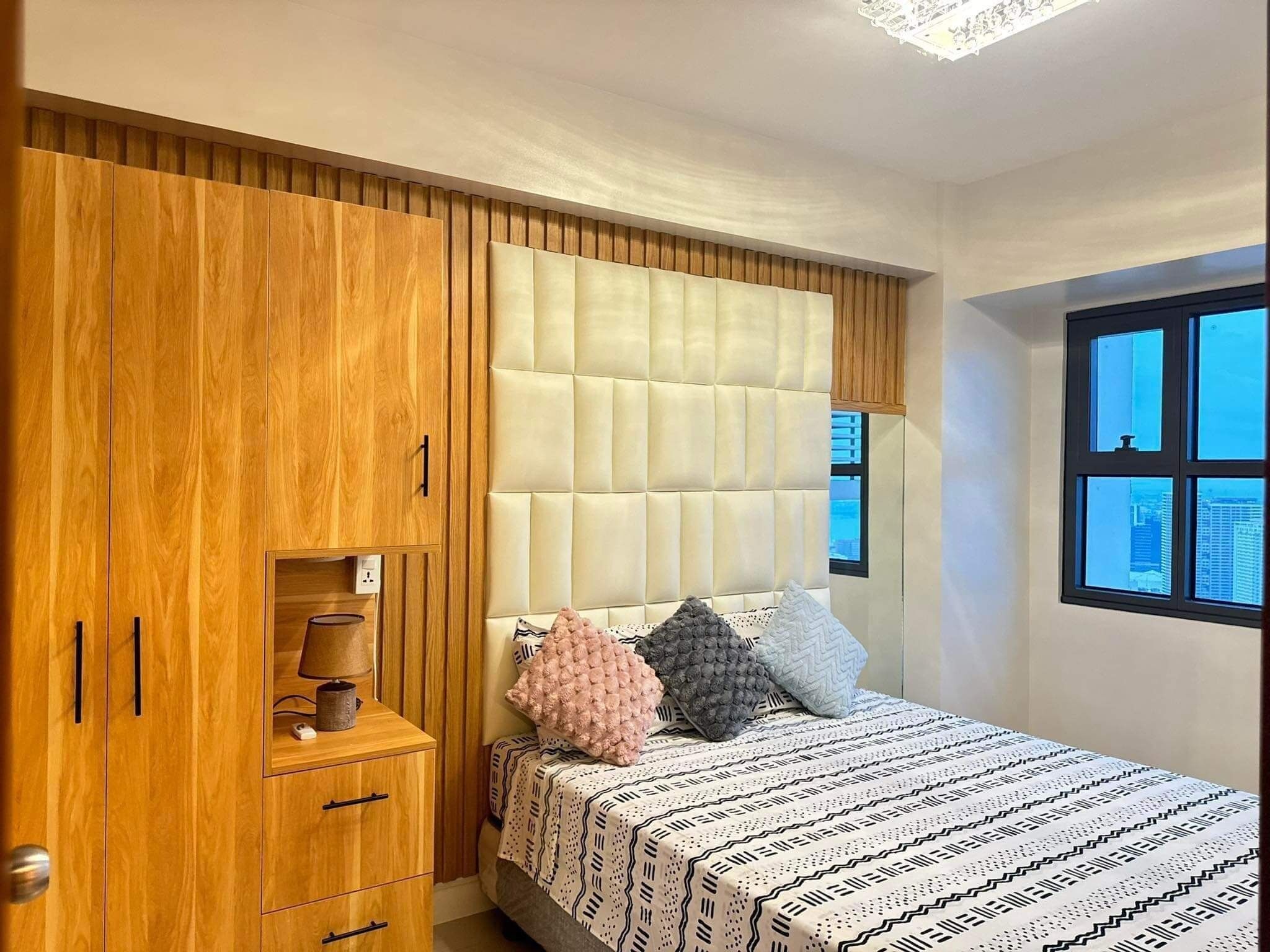 A bedroom with a wooden wardrobe, a cushioned headboard, a bed with patterned bedding, and a window with a city view.